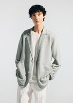 Wearing the TYPE-K 008 by A-POC ABLE ISSEY MIYAKE, a person with short, curly hair poses against a plain light background in a light gray, wrinkle-resistant blazer over a sheer white shirt and white pinstriped pants.