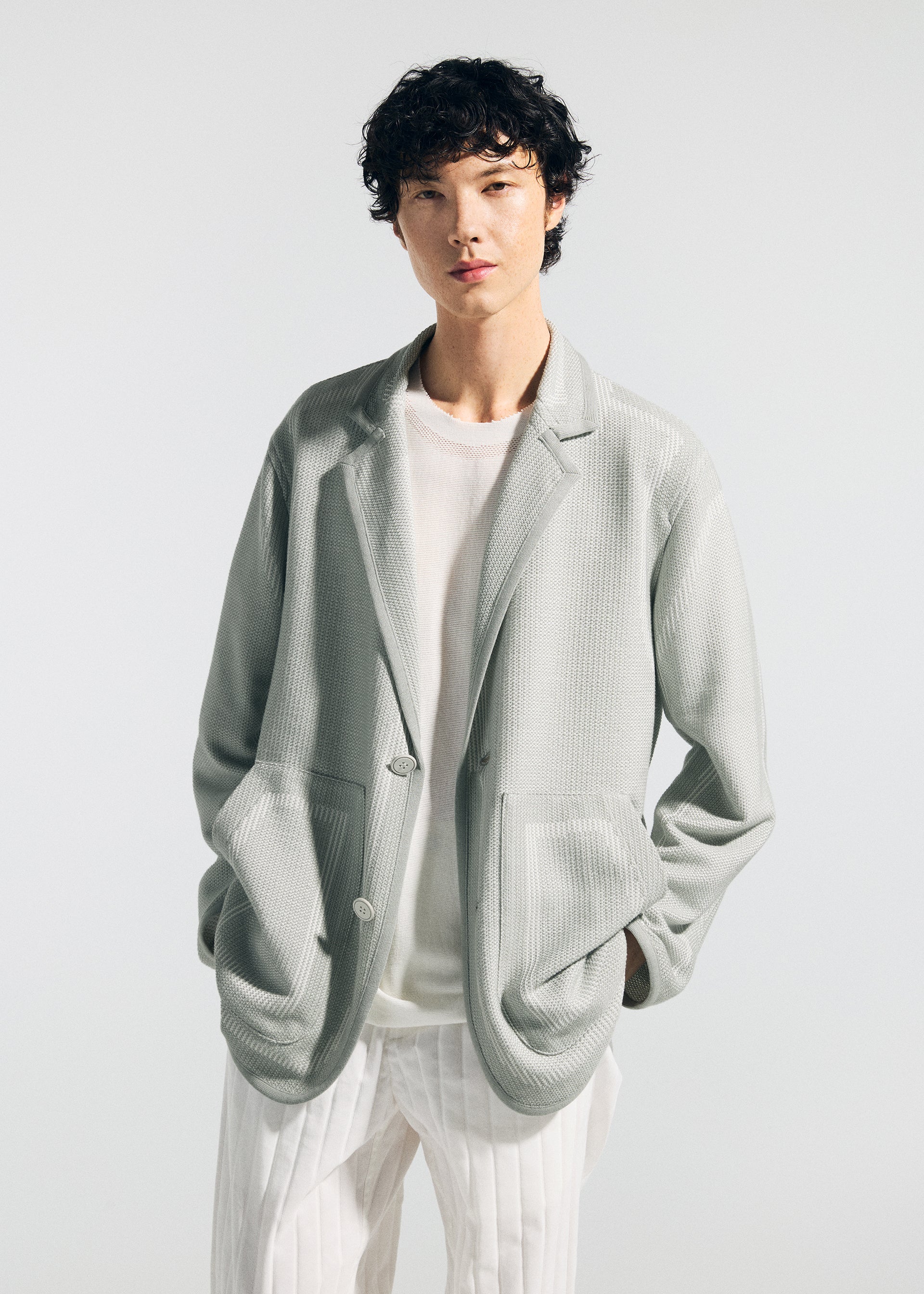 Zoomed view Wearing the TYPE-K 008 by A-POC ABLE ISSEY MIYAKE, a person with short, curly hair poses against a plain light background in a light gray, wrinkle-resistant blazer over a sheer white shirt and white pinstriped pants.