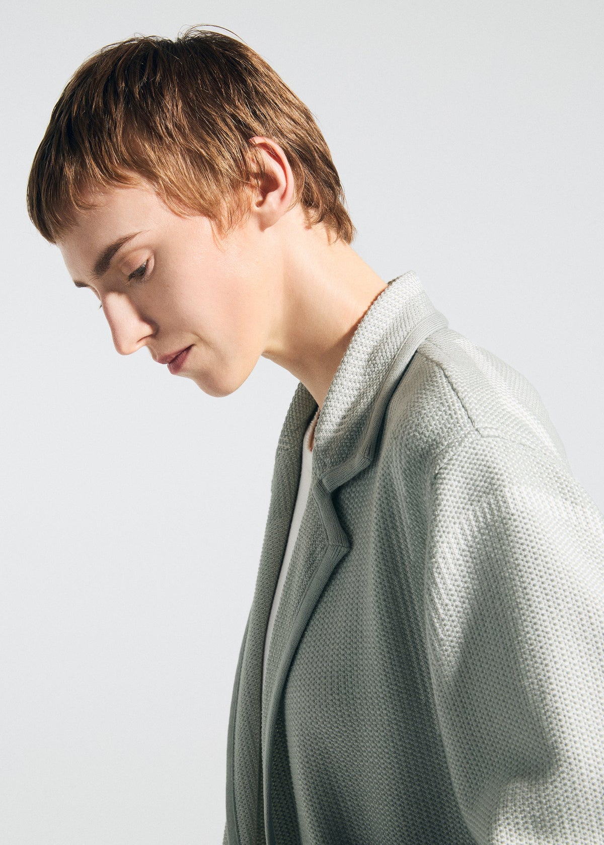 A person with short, light brown hair in profile, looking down calmly, wears the TYPE-K 008 jacket by A-POC ABLE ISSEY MIYAKE over a white top against a plain, light background. view 10 of 11
