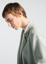 A person with short, light brown hair in profile, looking down calmly, wears the TYPE-K 008 jacket by A-POC ABLE ISSEY MIYAKE over a white top against a plain, light background.