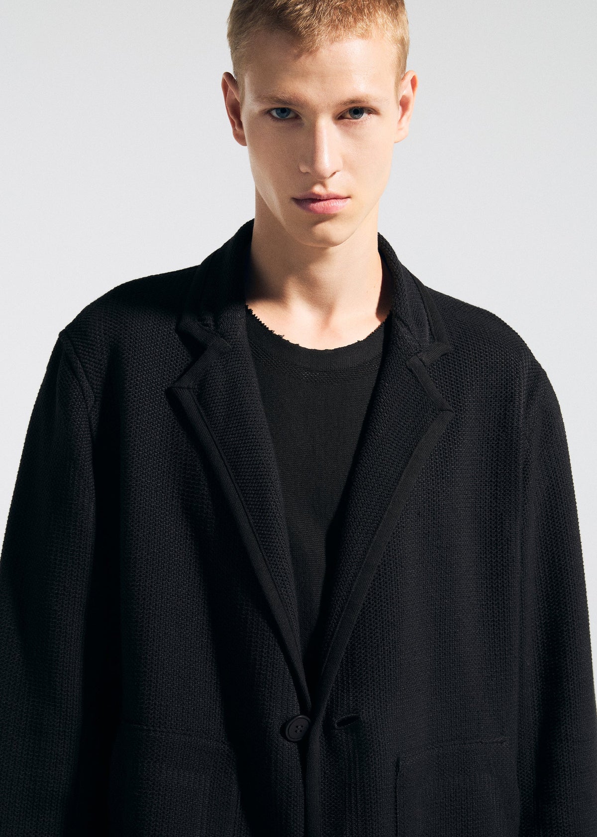 Wearing the TYPE-K 008 wrinkle-resistant black blazer by A-POC ABLE ISSEY MIYAKE over a black shirt, a short blonde-haired person stands against a plain light background, looking directly at the camera with a neutral expression. view 11 of 11