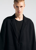 Wearing the TYPE-K 008 wrinkle-resistant black blazer by A-POC ABLE ISSEY MIYAKE over a black shirt, a short blonde-haired person stands against a plain light background, looking directly at the camera with a neutral expression.