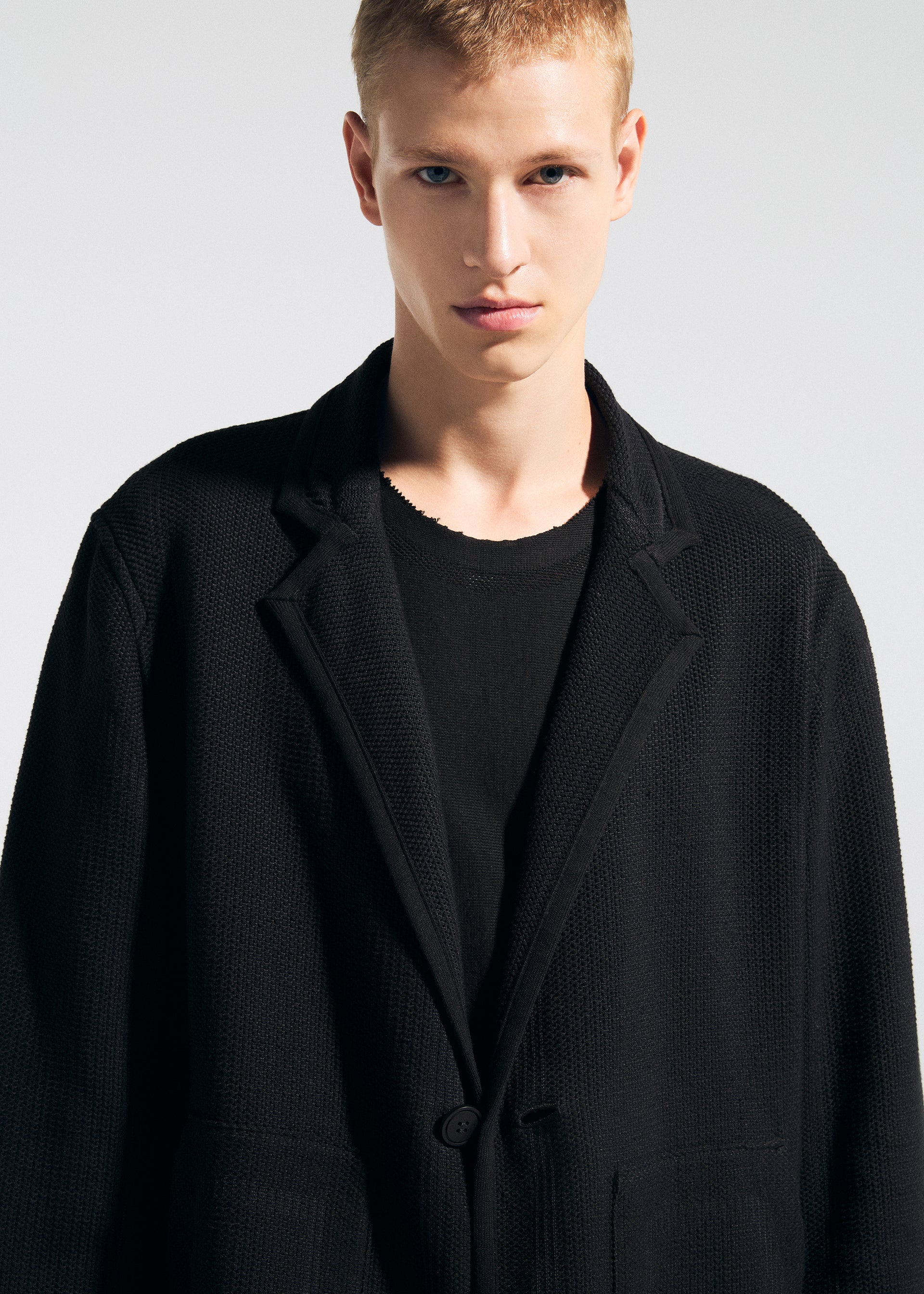 Zoomed view Wearing the TYPE-K 008 wrinkle-resistant black blazer by A-POC ABLE ISSEY MIYAKE over a black shirt, a short blonde-haired person stands against a plain light background, looking directly at the camera with a neutral expression.