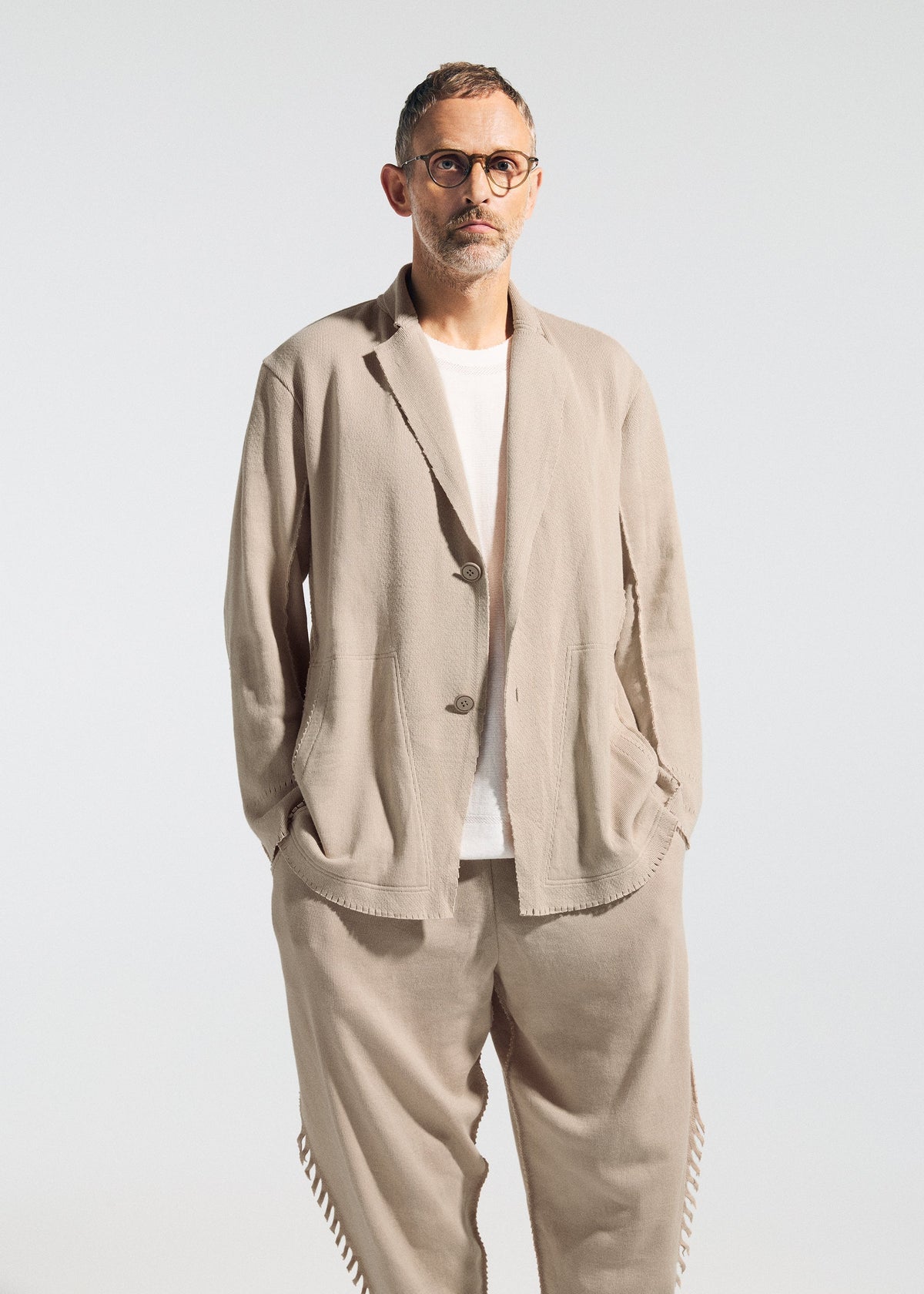 A man with short hair, glasses, and a beard wears the TYPE-A 003 by A-POC ABLE ISSEY MIYAKE—a relaxed beige suit styled over a white t-shirt and knit tapered pants—standing hands-in-pockets against a plain light background. view 7 of 7