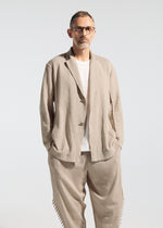 A man with short hair, glasses, and a beard wears the TYPE-A 003 by A-POC ABLE ISSEY MIYAKE—a relaxed beige suit styled over a white t-shirt and knit tapered pants—standing hands-in-pockets against a plain light background.