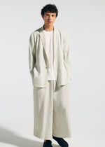 A person with short curly hair wears the TYPE-A 003 suit by A-POC ABLE ISSEY MIYAKE—loose, off-white knit wide pants and matching blazer over a white shirt, paired with black shoes. They stand against a plain light background.