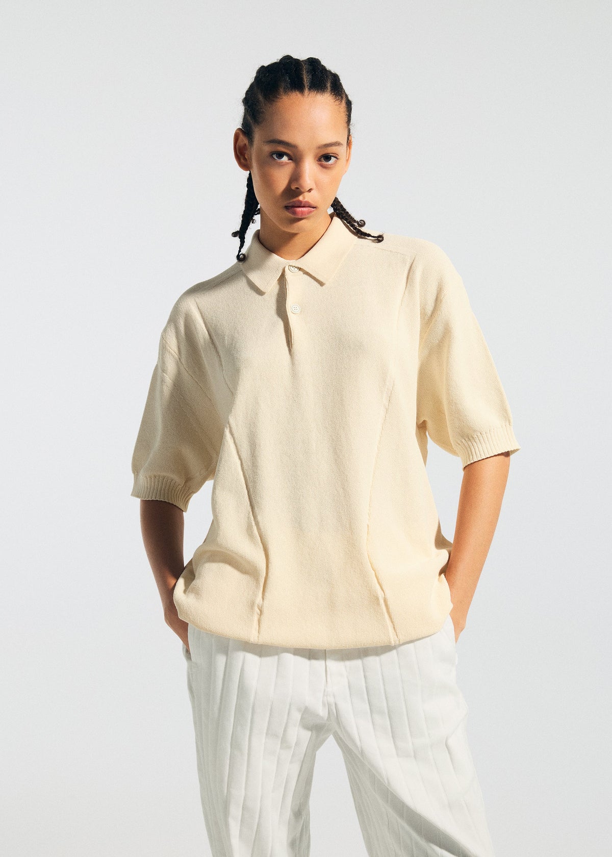 Wearing the TYPE-K 003-2 cream short-sleeved collared shirt by A-POC ABLE ISSEY MIYAKE and white pinstriped pants, a person with braided hair stands against a light background with hands in pockets. view 8 of 9