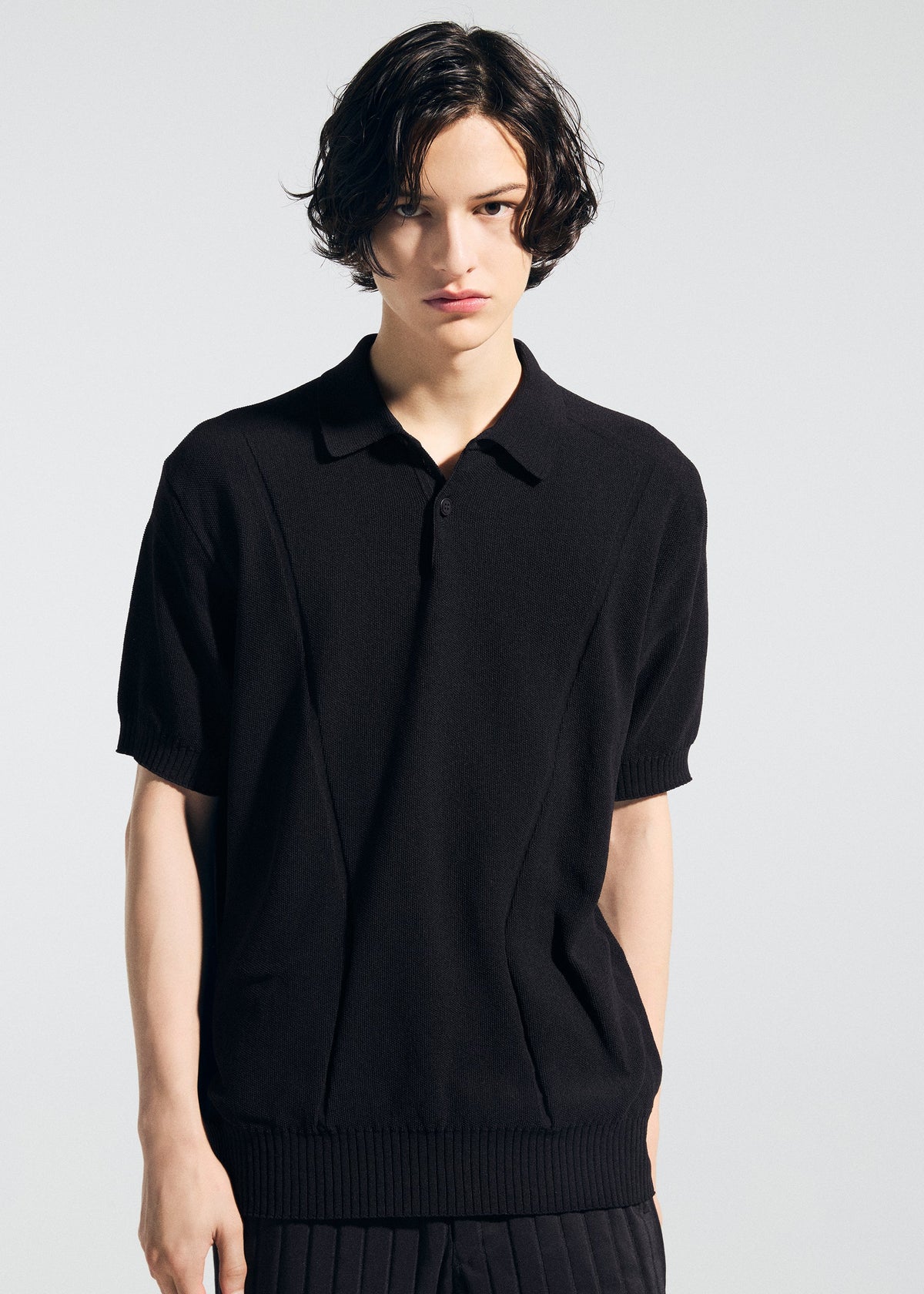A person with short, wavy dark hair wears the A-POC ABLE ISSEY MIYAKE TYPE-K 003-2 black knit polo shirt and black pants, standing against a plain light gray background and looking directly at the camera with a neutral expression. view 9 of 9