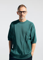 A man with short light hair, a beard, and black-rimmed glasses faces forward wearing the A-POC ABLE ISSEY MIYAKE TYPE-K 003-1—a stretchy, comfortable short-sleeved knit top—with dark pants and his hands in his pockets against a plain light background.