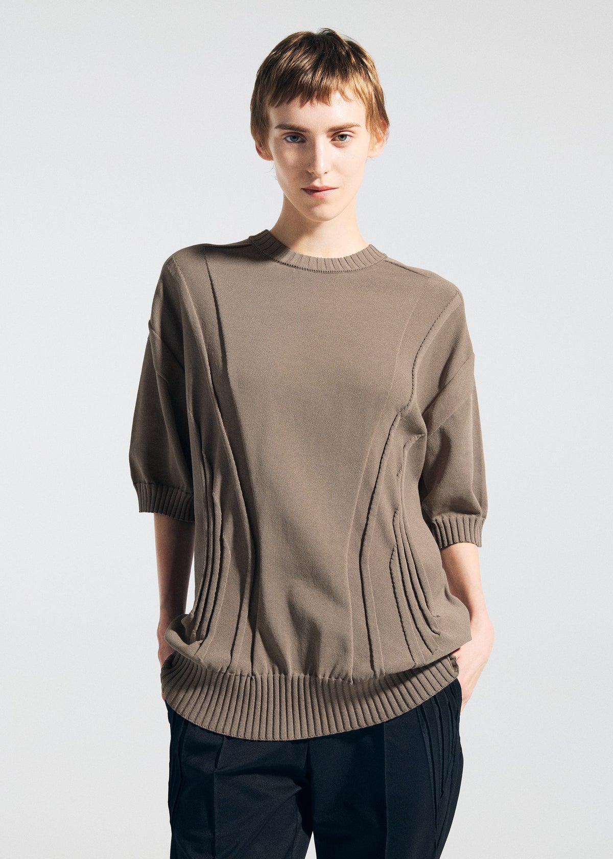 Wearing the TYPE-K 003-1 taupe weft knit top by A-POC ABLE ISSEY MIYAKE, a person with short light brown hair poses in black pants against a plain light background, hands in pockets. view 5 of 7