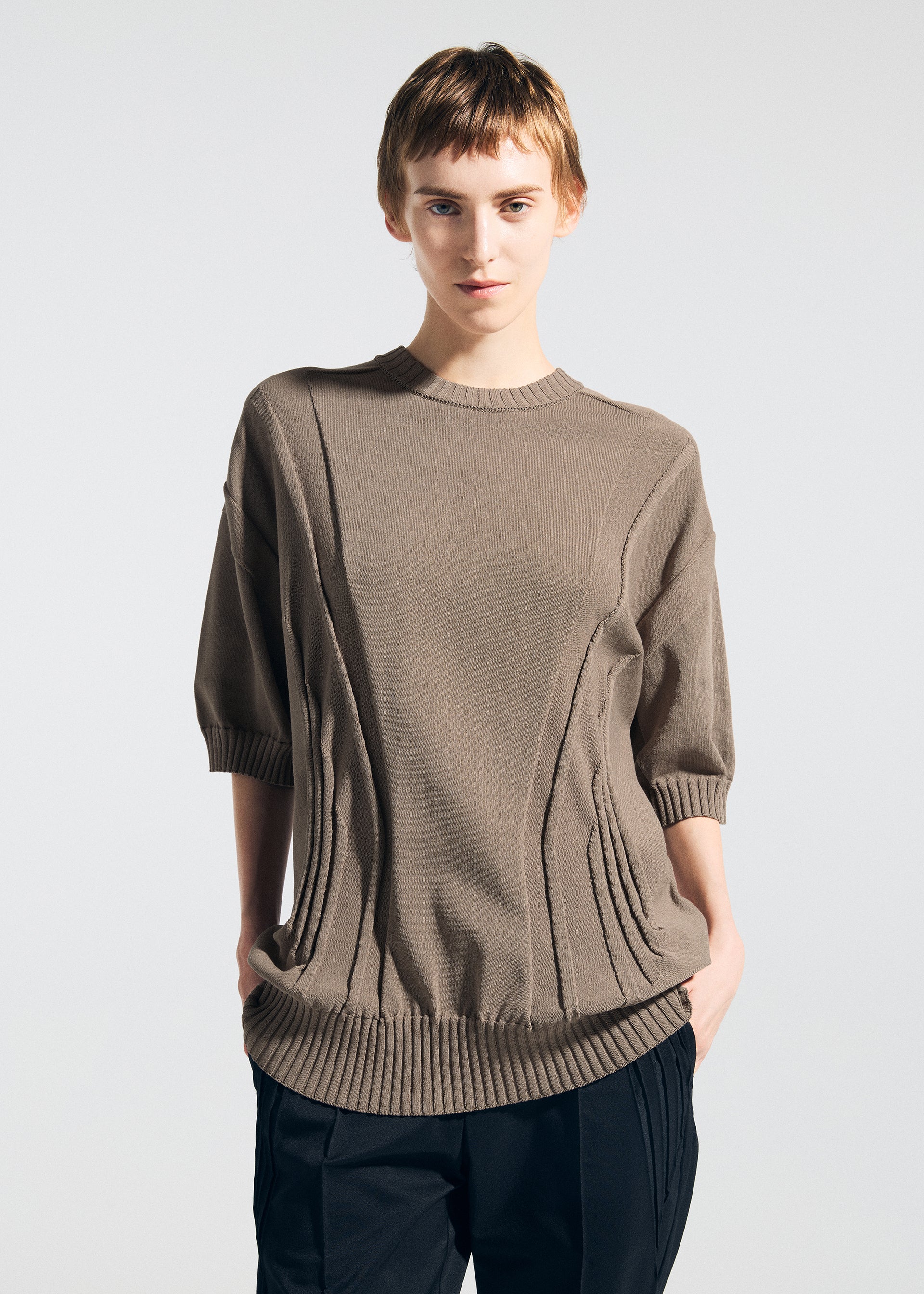 Zoomed view A person with short light brown hair models the TYPE-K 003-1 sweater by A-POC ABLE ISSEY MIYAKE, featuring ribbed cuffs and hem, paired with black pants. They stand against a light background, hands in pockets, facing the camera.