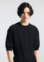A person with wavy, chin-length dark hair wears the TYPE-K 003-1 black short-sleeved knit top by A-POC ABLE ISSEY MIYAKE, featuring ribbed edges and weft knitting, standing against a plain light background and looking slightly to the side.