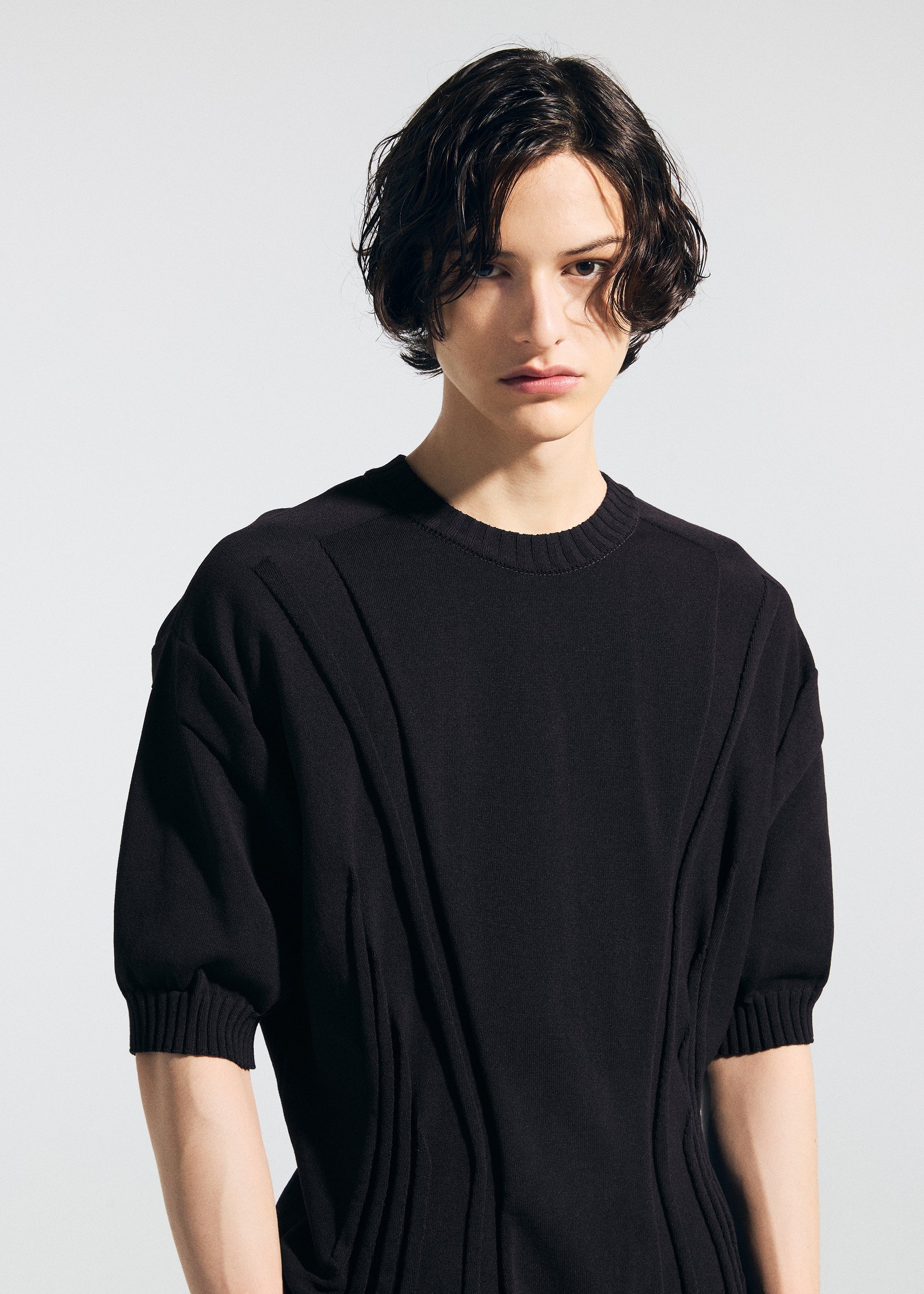 Zoomed view A person with wavy, chin-length dark hair wears the A-POC ABLE ISSEY MIYAKE TYPE-K 003-1 black short-sleeve knit top made with weft knitting, looking towards the camera against a plain light background.