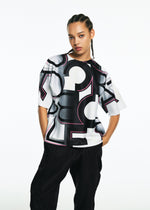 A person with braided hair wears the TYPE-XV SOAR NY project 002 shirt by A-POC ABLE ISSEY MIYAKE in a bold black, white, and magenta geometric pattern, paired with black pants, standing against a white background with hands in pockets.