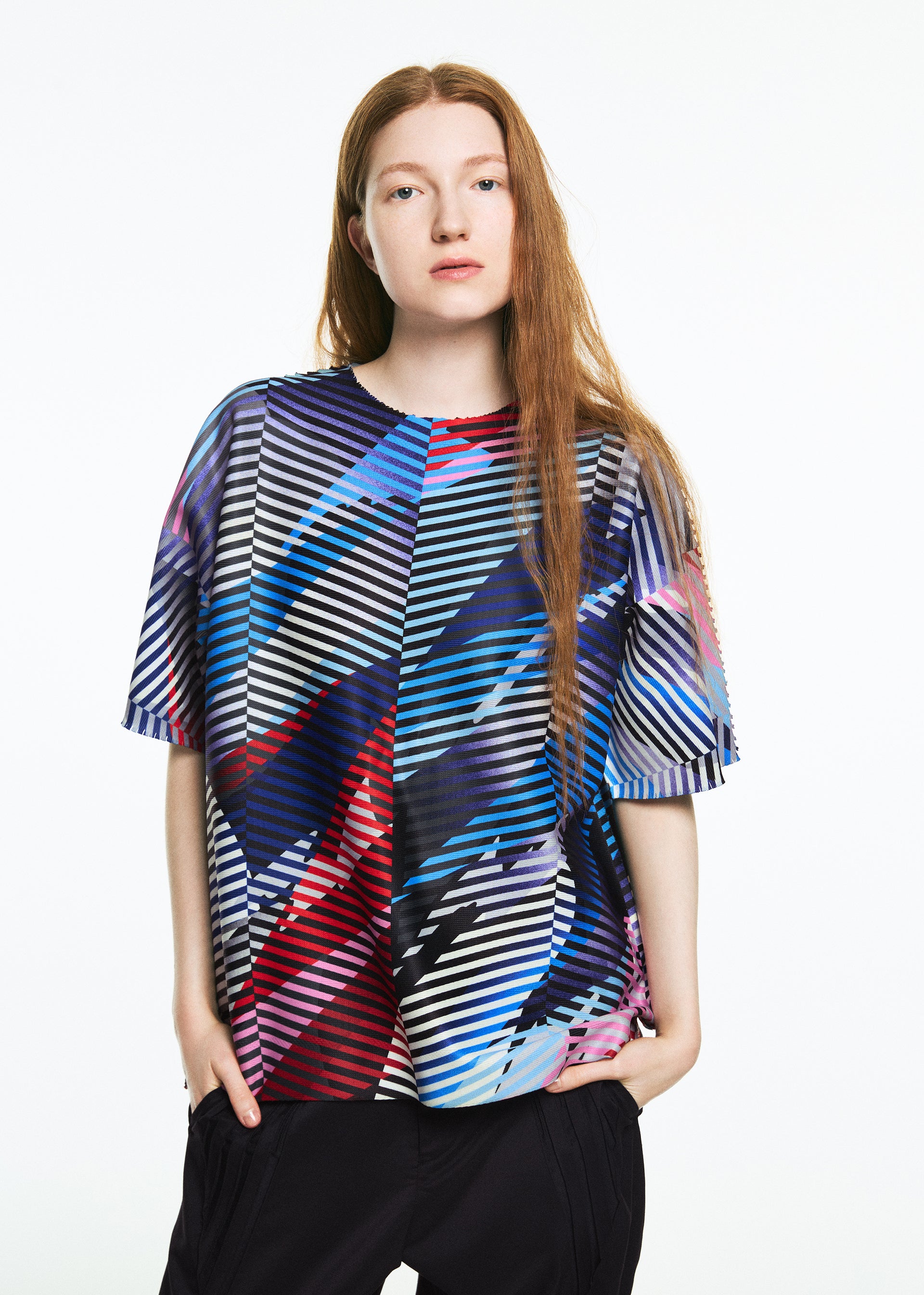 Zoomed view A person with long, straight reddish-brown hair wears the TYPE-XV SOAR NY project 003 short-sleeve knit top by A-POC ABLE ISSEY MIYAKE, featuring bold colorful geometric stripes, paired with black pants against a plain white background.