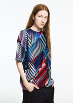 A person with long, straight, reddish-brown hair wears the TYPE-XV SOAR NY project 003 short-sleeve knit top by A-POC ABLE ISSEY MIYAKE, featuring colorful geometric diagonal stripes, and dark pants against a plain white background.