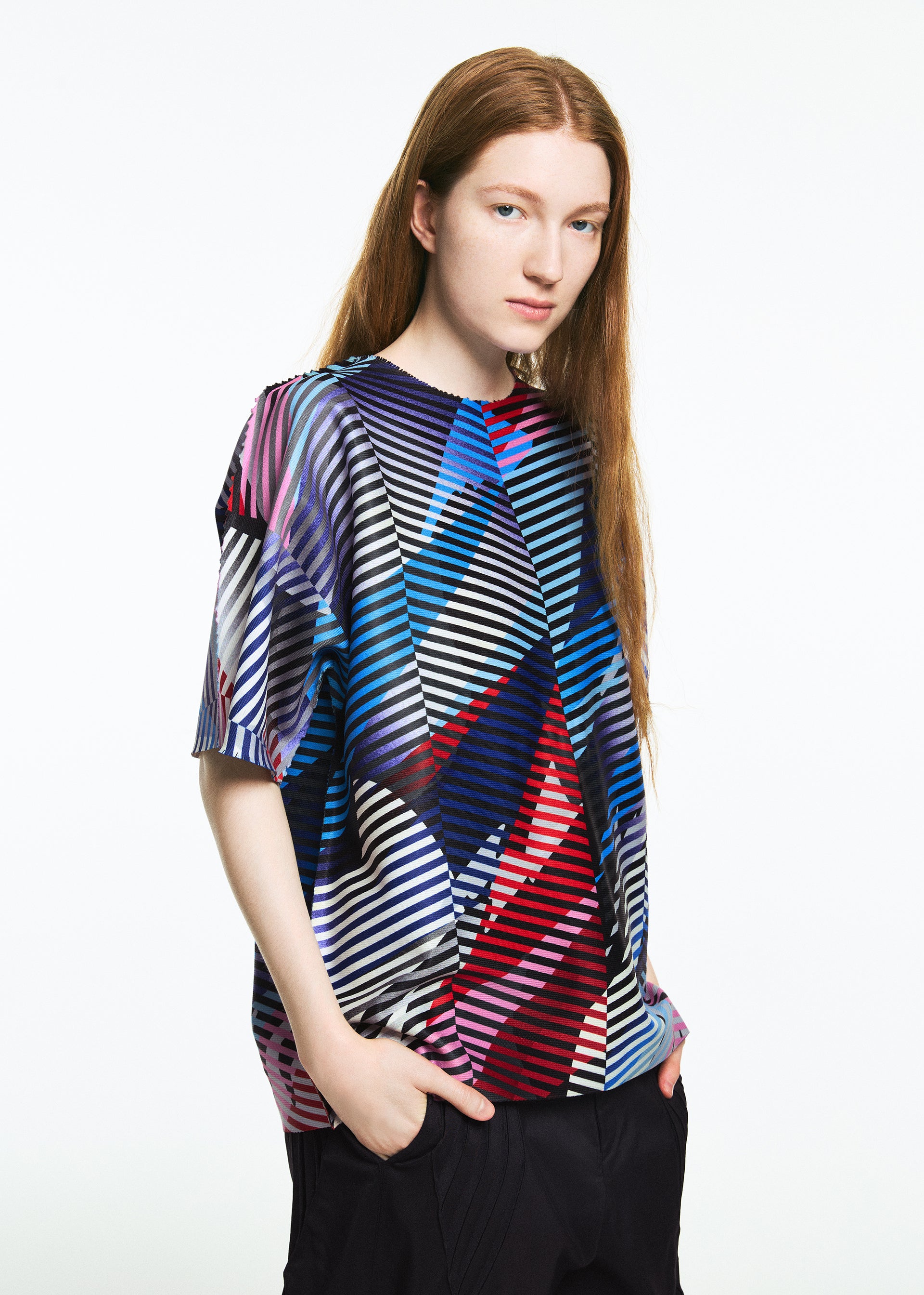 Zoomed view A person with long, straight, reddish-brown hair wears the TYPE-XV SOAR NY project 003 short-sleeve knit top by A-POC ABLE ISSEY MIYAKE, featuring colorful geometric diagonal stripes, and dark pants against a plain white background.