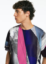 A young person with short curly hair looks left, wearing the TYPE-XV SOAR NY project 003 short-sleeve knit top by A-POC ABLE ISSEY MIYAKE, featuring abstract geometric patterns in pink, blue, black, white, and gray on a plain white background.