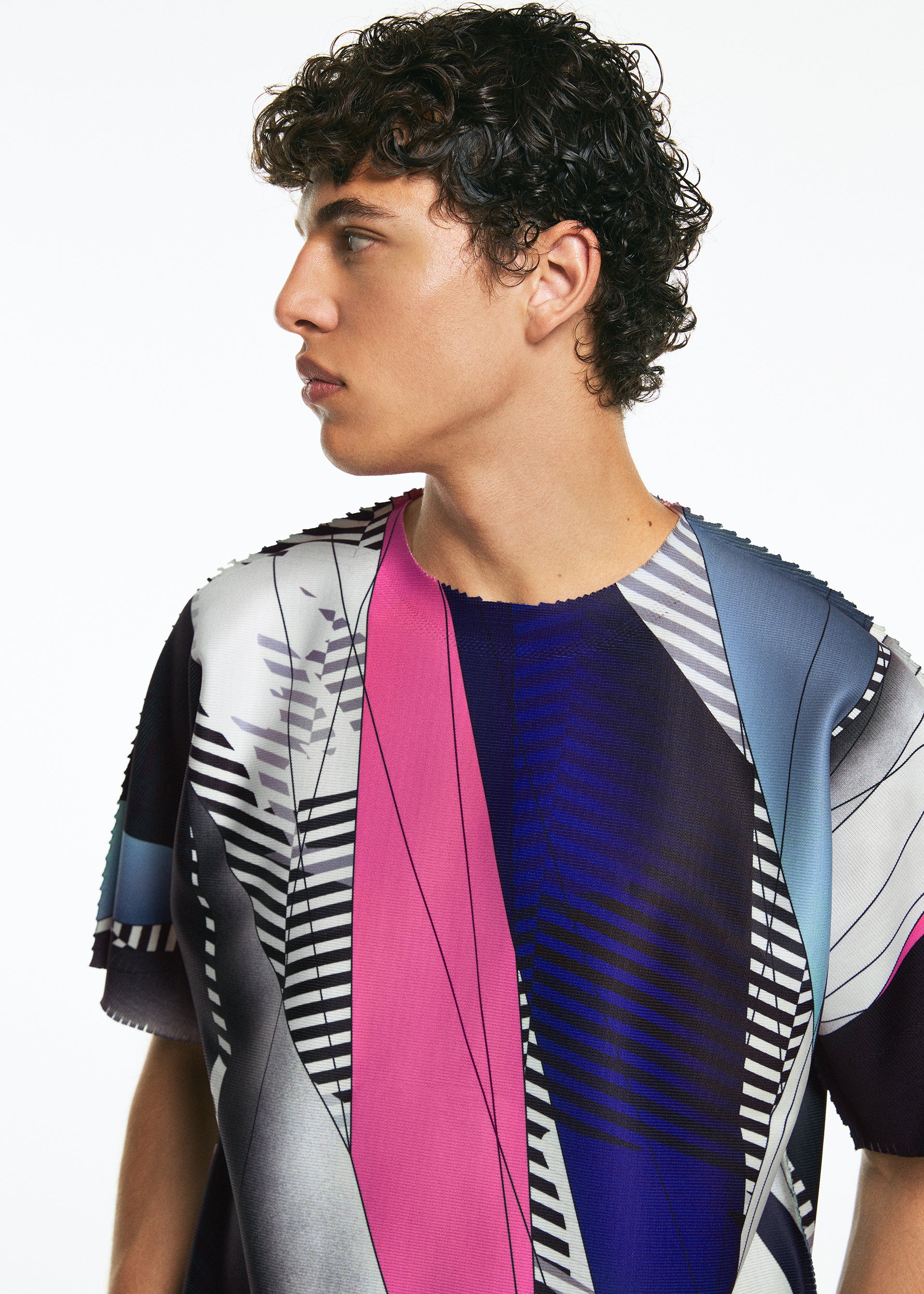 Zoomed view A young person with short curly hair looks left, wearing the TYPE-XV SOAR NY project 003 short-sleeve knit top by A-POC ABLE ISSEY MIYAKE, featuring abstract geometric patterns in pink, blue, black, white, and gray on a plain white background.