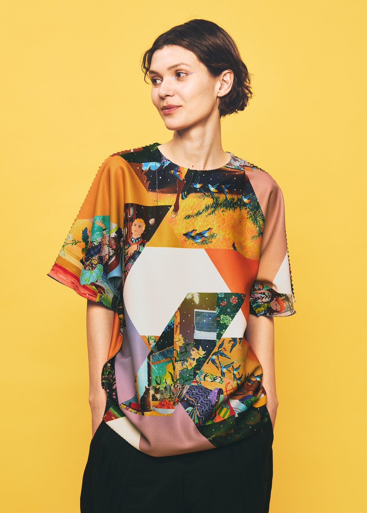 A person with short brown hair stands against a yellow background, wearing the TYPE-XII Tomokazu Matsuyama project 004 shirt from A-POC ABLE ISSEY MIYAKE, featuring colorful geometric and nature-inspired designs from the contemporary artist collaboration. Their hands are in their pockets as they look slightly to the side and smile softly. view 9 of 15