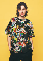 A person with wavy brown hair stands against a yellow background, confidently looking at the camera with hands in their pockets. They are wearing the TYPE-XII Tomokazu Matsuyama project 004 shirt—a colorful, abstract-patterned piece from the A-POC ABLE ISSEY MIYAKE collaboration with contemporary artist Tomokazu Matsuyama—paired with black pants.
