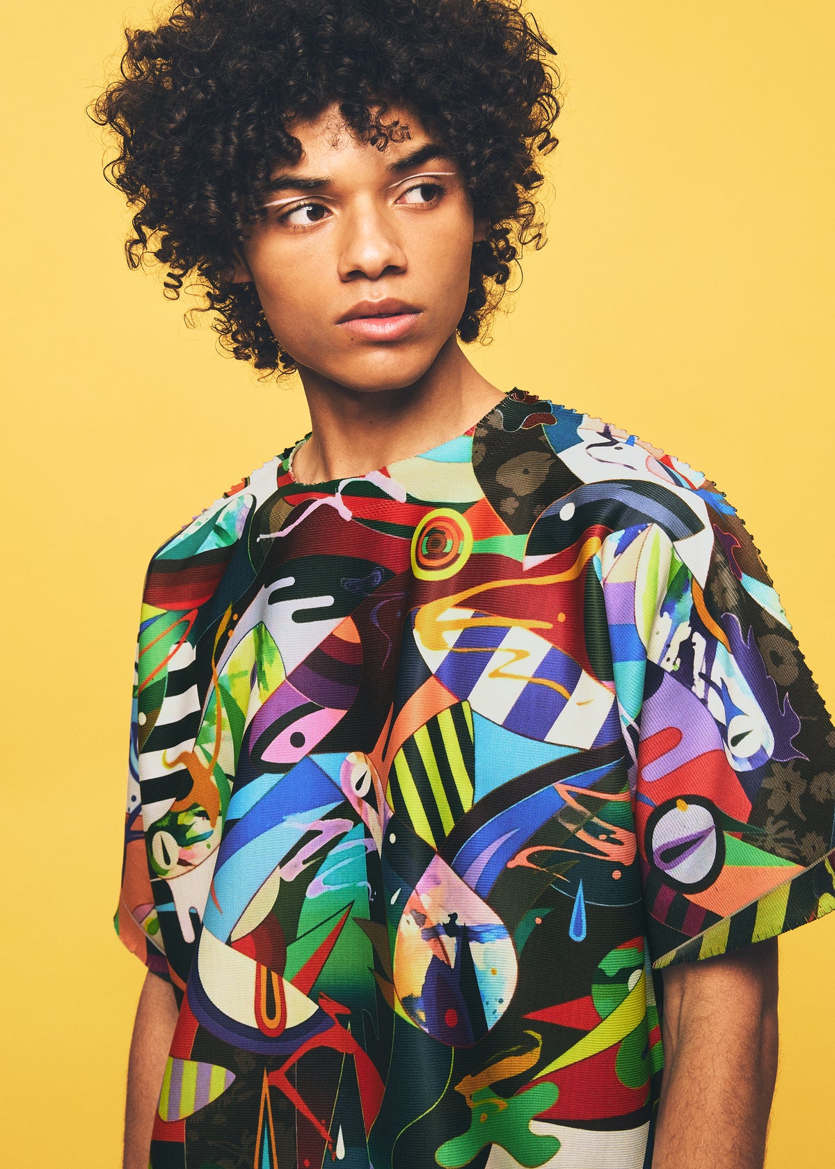A person with curly hair looks to the side, wearing the TYPE-XII Tomokazu Matsuyama project 004 top from A-POC ABLE ISSEY MIYAKE, featuring a colorful abstract pattern inspired by a contemporary artist collaboration, set against a solid yellow background. view 12 of 15