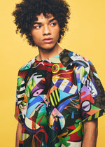 A person with curly hair looks to the side, wearing the TYPE-XII Tomokazu Matsuyama project 004 top from A-POC ABLE ISSEY MIYAKE, featuring a colorful abstract pattern inspired by a contemporary artist collaboration, set against a solid yellow background.