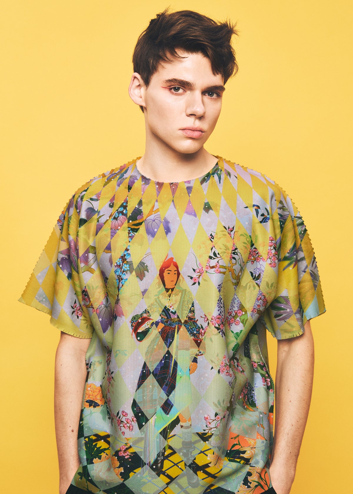 A young person with short dark hair wears the TYPE-XII Tomokazu Matsuyama project 004 shirt from A-POC ABLE ISSEY MIYAKE, a vibrant, geometric and character-patterned piece created in collaboration with contemporary artist Tomokazu Matsuyama—standing out against a vivid yellow background. view 13 of 15