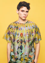 A young person with short dark hair wears the TYPE-XII Tomokazu Matsuyama project 004 shirt from A-POC ABLE ISSEY MIYAKE, a vibrant, geometric and character-patterned piece created in collaboration with contemporary artist Tomokazu Matsuyama—standing out against a vivid yellow background.
