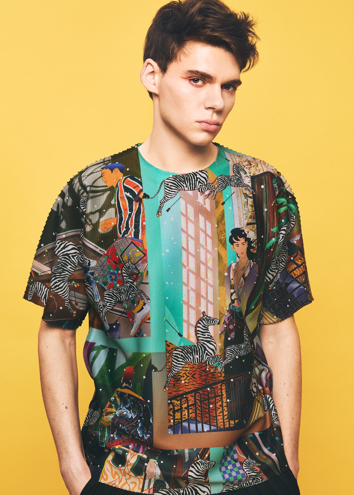 A young person stands against a yellow background, wearing the TYPE-XII Tomokazu Matsuyama project 004 t-shirt from A-POC ABLE ISSEY MIYAKE. The colorful graphic tee, created in collaboration with contemporary artist Tomokazu Matsuyama, features illustrated zebras, cityscapes, and various artistic designs. They look directly at the camera with a neutral expression. view 14 of 15