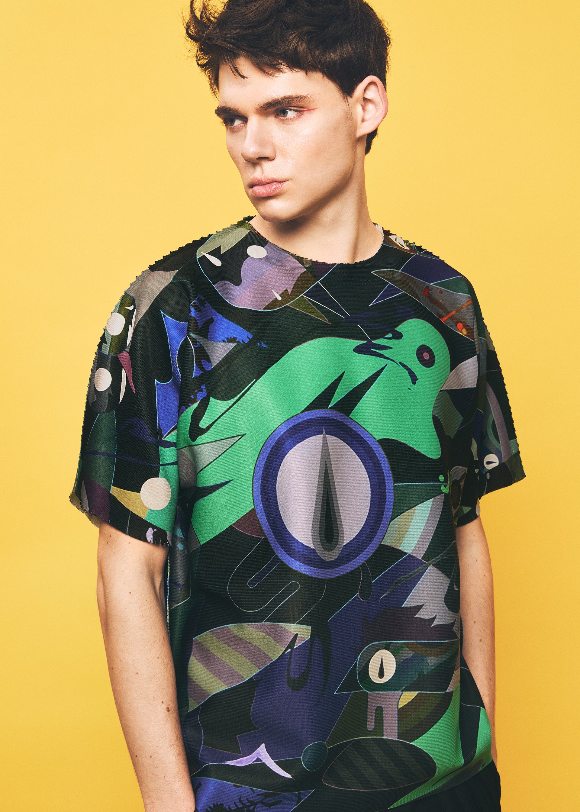 Zoomed view A young person with short brown hair wears the TYPE-XII Tomokazu Matsuyama project 004 shirt by A-POC ABLE ISSEY MIYAKE, featuring a colorful abstract pattern with green and black shapes, reminiscent of a contemporary artist collaboration, standing against a solid yellow background.