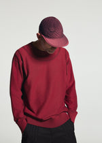 Wearing the TYPE-O CAP by A-POC ABLE ISSEY MIYAKE and a matching red sweater, a person stands against a plain background, looking down with hands in their pockets.
