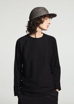 A person with curly hair wears the TYPE-O CAP by A-POC ABLE ISSEY MIYAKE, paired with a black long-sleeve shirt and dark pants, looking sideways against a plain light background.