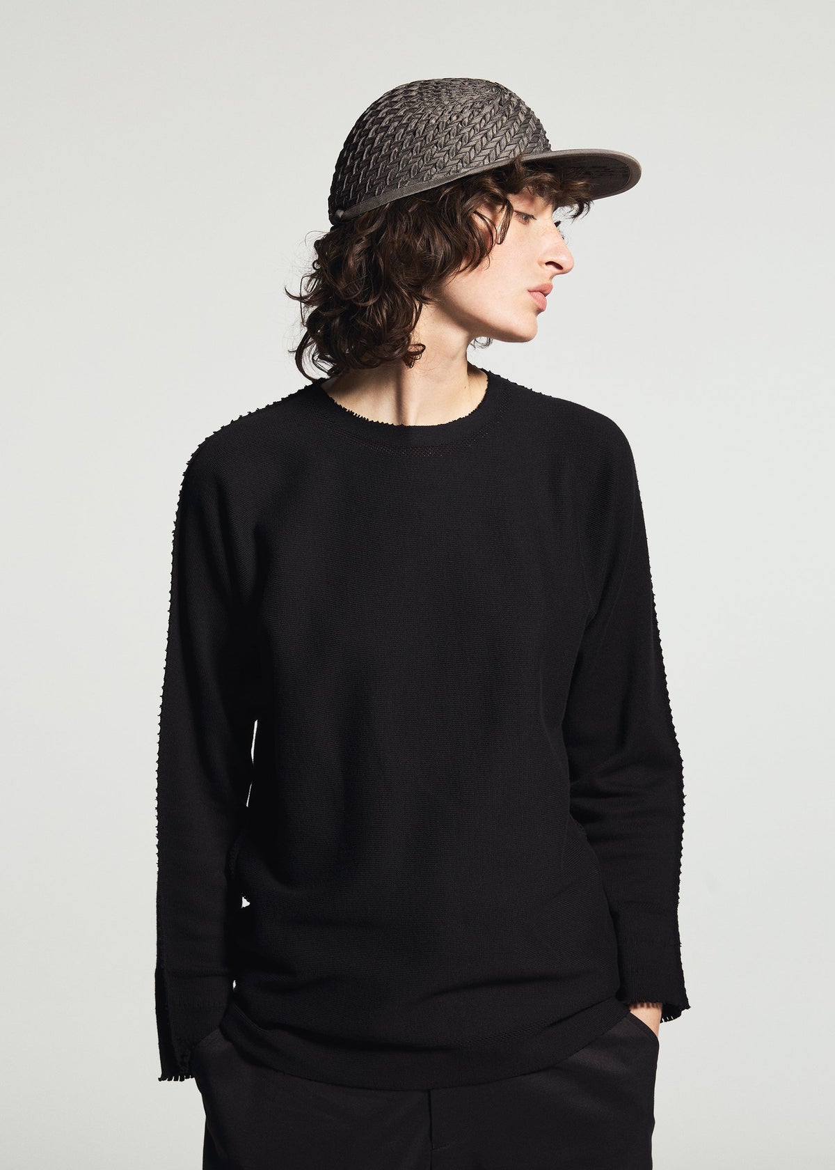 Zoomed view A person with curly hair wears the TYPE-O CAP by A-POC ABLE ISSEY MIYAKE, paired with a black long-sleeve shirt and dark pants, looking sideways against a plain light background.