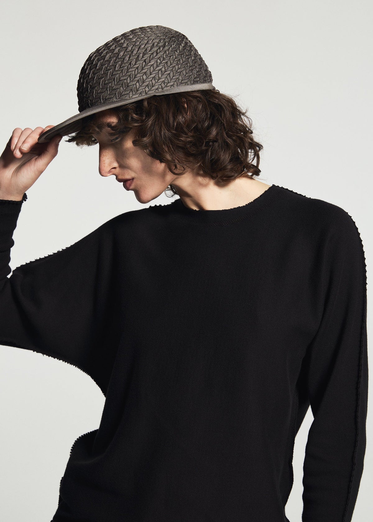 Person with curly brown hair wearing the TYPE-O CAP by A-POC ABLE ISSEY MIYAKE and a black long-sleeve top, tilting the cap down, looking slightly downward against a plain light background. view 13 of 16