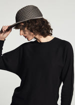 Person with curly brown hair wearing the TYPE-O CAP by A-POC ABLE ISSEY MIYAKE and a black long-sleeve top, tilting the cap down, looking slightly downward against a plain light background.