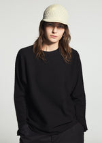 A person with long brown hair wears the TYPE-O CAP by A-POC ABLE ISSEY MIYAKE and a loose black sweater, standing against a plain light background with a neutral expression.