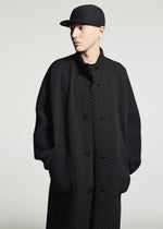A person in a textured black coat and the TYPE-O CAP by A-POC ABLE ISSEY MIYAKE stands with hands in pockets, gazing to the side against a plain light background.