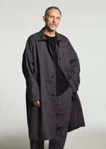 A man with short hair and a beard wears the TYPE-U 001-1 coat by A-POC ABLE ISSEY MIYAKE—a long, dark gray, wrinkle-resistant garment with adjustable cuffs—layered over a black shirt and pants, standing against a plain light background with a neutral expression.