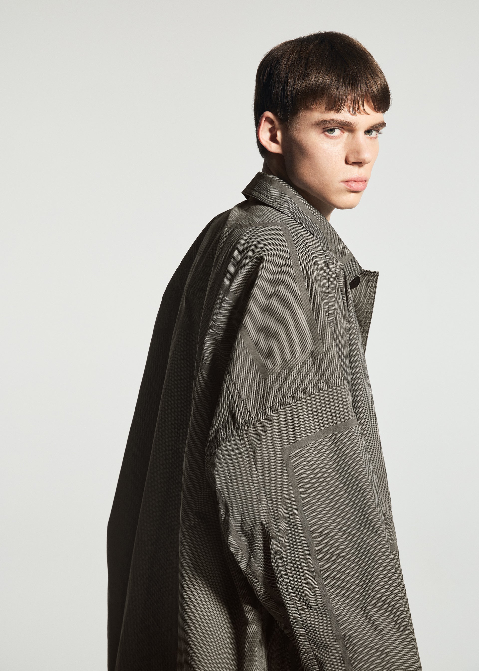 Zoomed view A young person with short brown hair wears the oversized, wrinkle-resistant TYPE-U 001-1 coat from A-POC ABLE ISSEY MIYAKE, glancing over their shoulder against a plain light background.