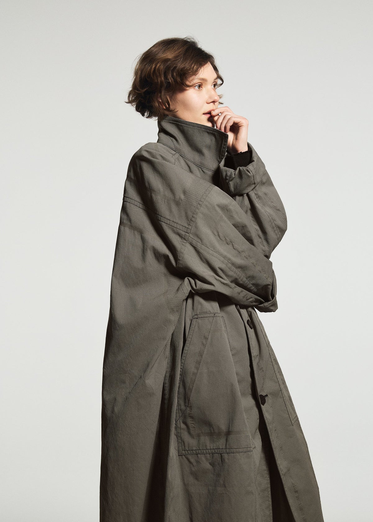 A person with short brown hair, dressed in the TYPE-U 001-1 shape-memory coat by A-POC ABLE ISSEY MIYAKE, stands in profile against a plain light background, thoughtfully touching their face with one hand. The long, oversized coat features adjustable cuffs and a wrinkle-resistant finish, offering both style and practicality to the scene. view 8 of 10
