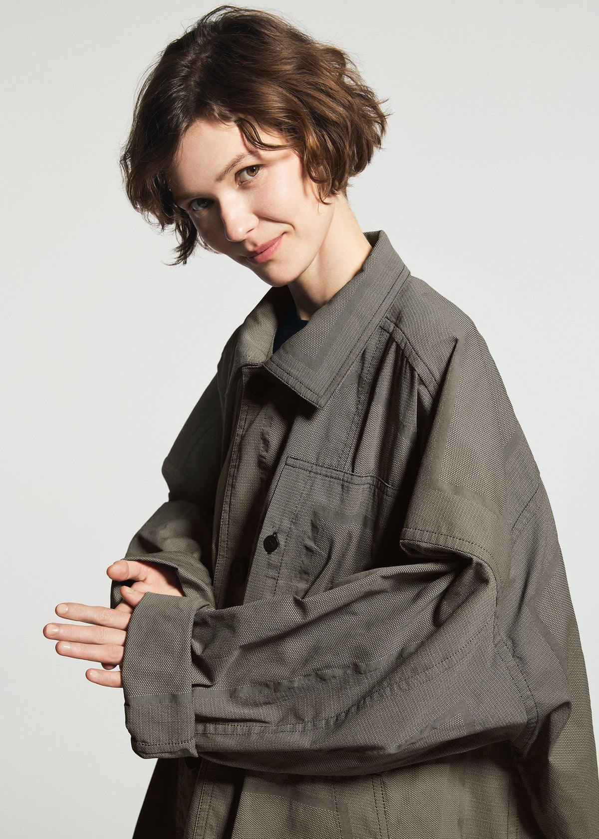 A person with short brown hair smiles, wearing the oversized TYPE-U 001-1 gray shape-memory coat with adjustable cuffs by A-POC ABLE ISSEY MIYAKE, standing against a plain light background. view 9 of 10