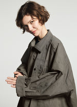 A person with short brown hair smiles, wearing the oversized TYPE-U 001-1 gray shape-memory coat with adjustable cuffs by A-POC ABLE ISSEY MIYAKE, standing against a plain light background.