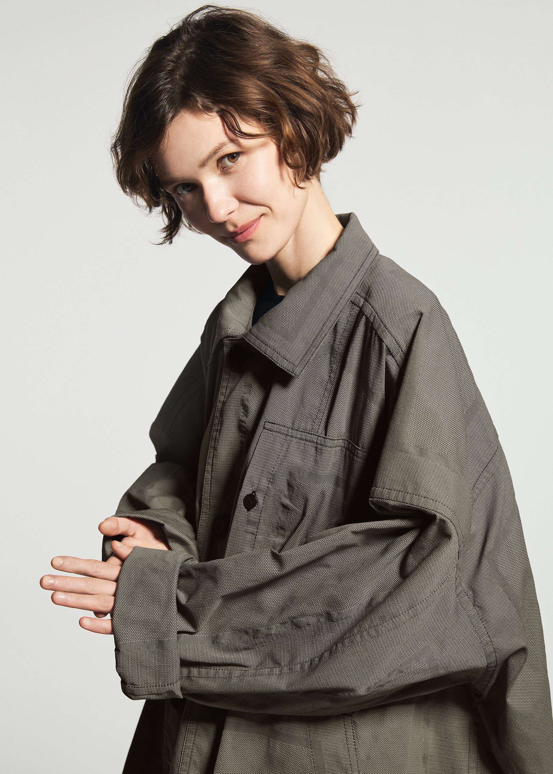 Zoomed view A person with short brown hair smiles, wearing the oversized TYPE-U 001-1 gray shape-memory coat with adjustable cuffs by A-POC ABLE ISSEY MIYAKE, standing against a plain light background.
