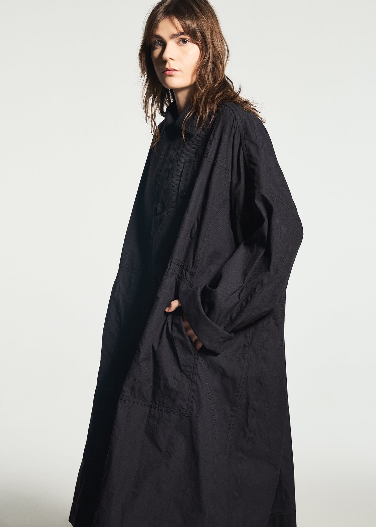 A person wearing the TYPE-U 001-1 long, loose-fitting black coat by A-POC ABLE ISSEY MIYAKE, featuring adjustable cuffs, stands against a plain light background, looking toward the camera with a neutral expression and one hand in their pocket. view 10 of 10