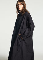 A person wearing the TYPE-U 001-1 long, loose-fitting black coat by A-POC ABLE ISSEY MIYAKE, featuring adjustable cuffs, stands against a plain light background, looking toward the camera with a neutral expression and one hand in their pocket.
