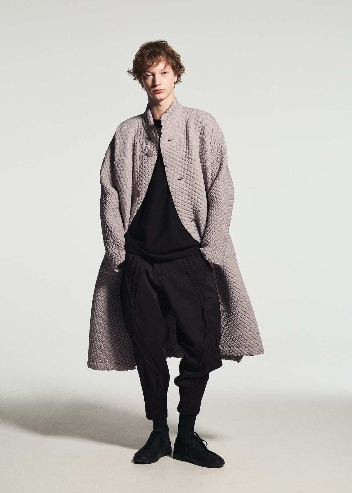 A person wears the TYPE-O 024 long coat by A-POC ABLE ISSEY MIYAKE in textured light gray, styled over a black top and pants with black shoes, standing against a plain white background. view 6 of 15
