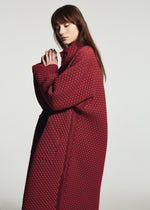 A woman with long brown hair wears the TYPE-O 024 by A-POC ABLE ISSEY MIYAKE, a textured oversized red coat, standing in profile against a plain light background with hands clasped near her face.