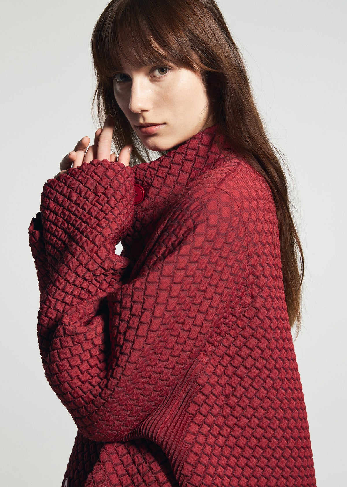 A woman with long brown hair wears the TYPE-O 024 textured red double-weave sweater by A-POC ABLE ISSEY MIYAKE, holding the sleeve close to her face against a plain background. view 9 of 15