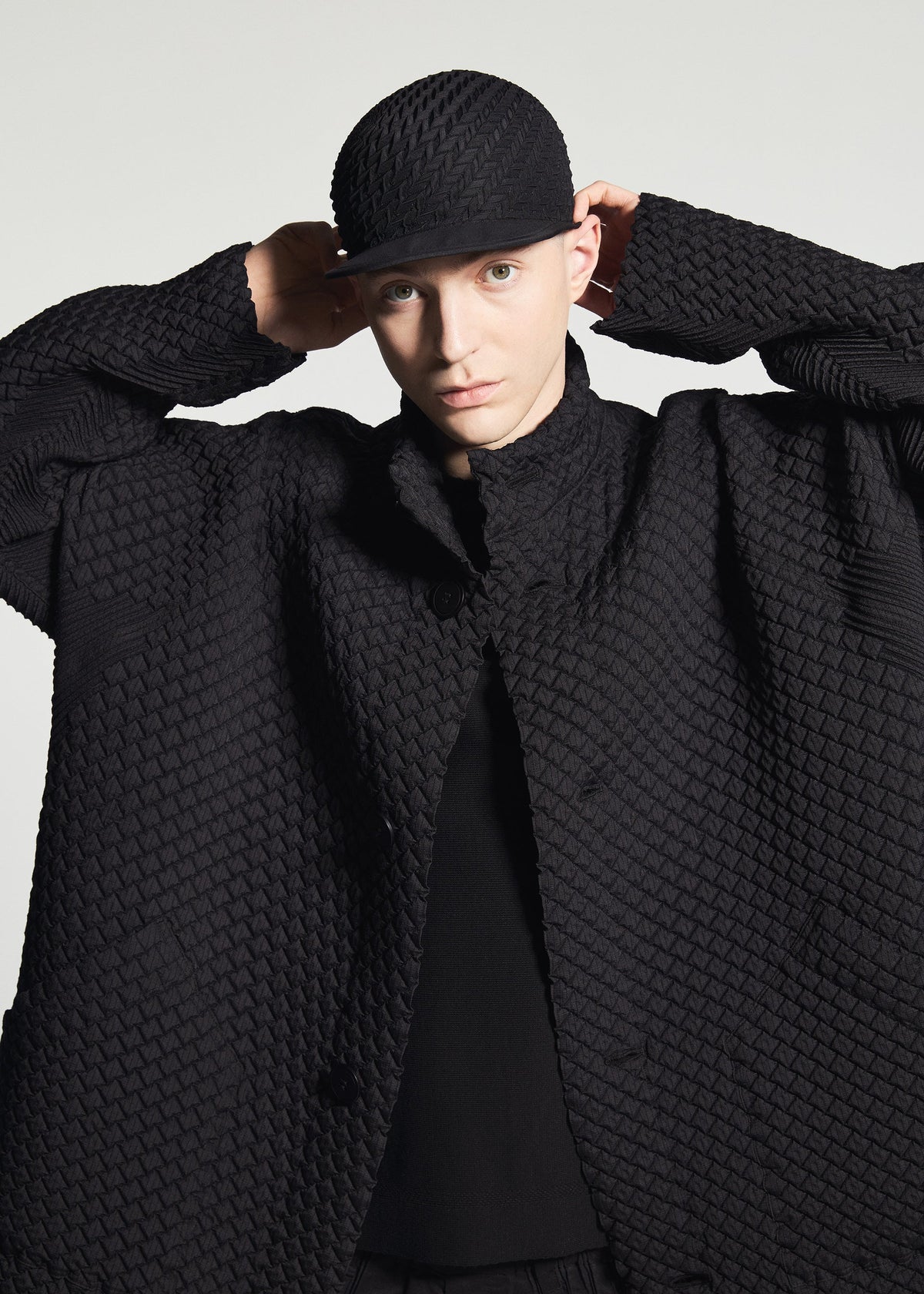Model wears TYPE-O 024 by A-POC ABLE ISSEY MIYAKE, a textured black coat in double-weave fabric with a matching cap, adjusting the hat while looking at the camera against a plain light background. view 11 of 15